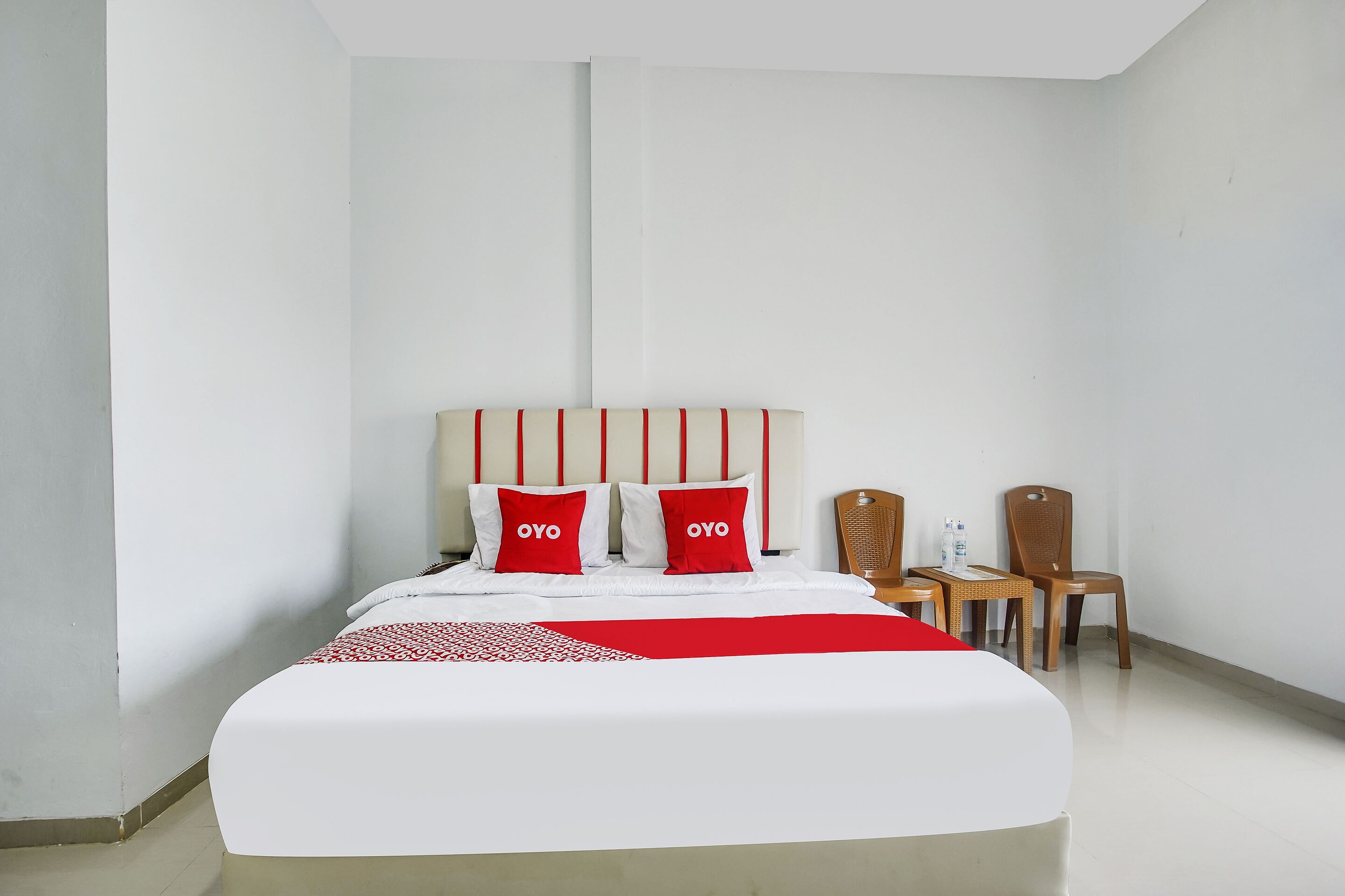 A hotel room with a large bed, two red pillows, a wooden chair, and a small table.