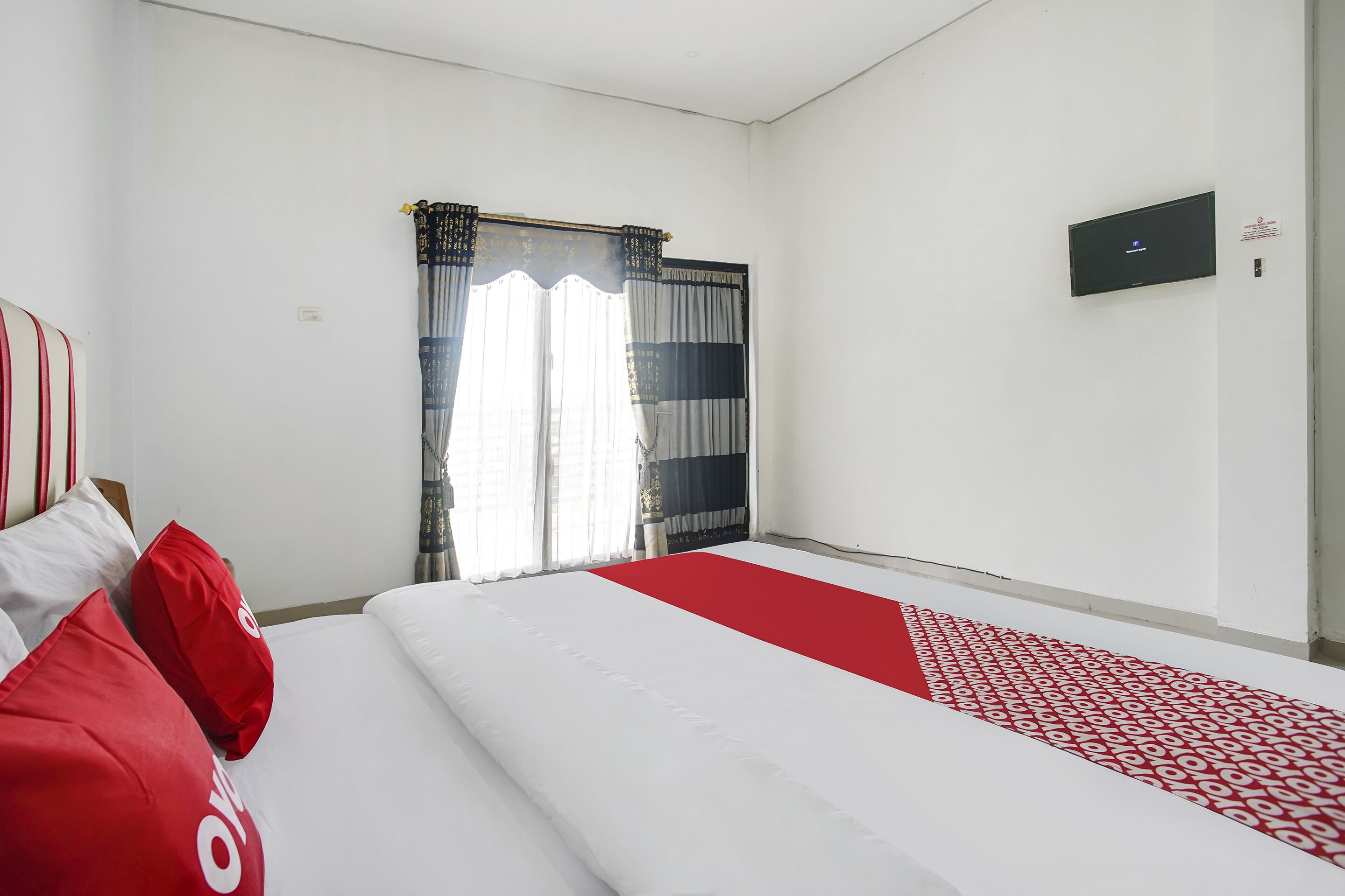A hotel room with a bed, a flat-screen TV mounted on the wall, and a window with curtains.