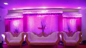 A stage with three ornate white sofas, a floral backdrop, and vertical light panels. - Hotel Shivam (Kalyan)