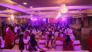 A large indoor event with rows of white chairs and tables, attendees in traditional attire, and chandeliers. - Hotel Shivam (Kalyan)