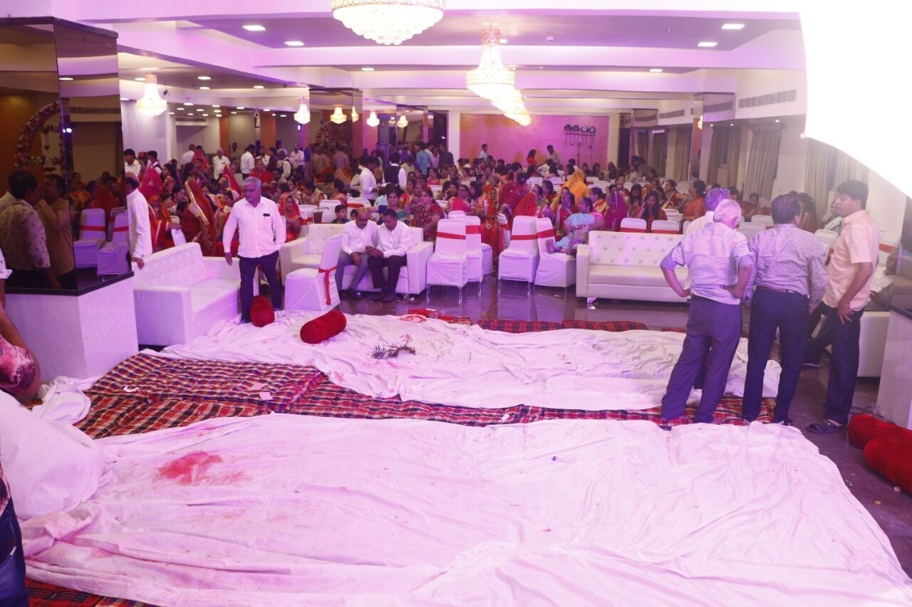 A large indoor event with a crowd seated on white chairs, a stage, and a large white cloth laid out on the floor.