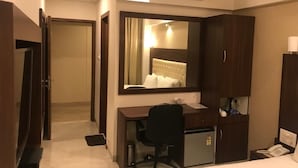 A hotel room with a desk, chair, and a large mirror. There is a bed with a white comforter, a bedside table with a lamp, and a phone. - Hotel Shivam (Kalyan)