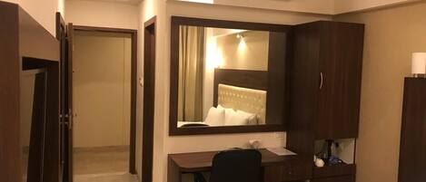 A hotel room with a desk, chair, and a large mirror. There is a bed with a white comforter, a bedside table with a lamp, and a phone.
