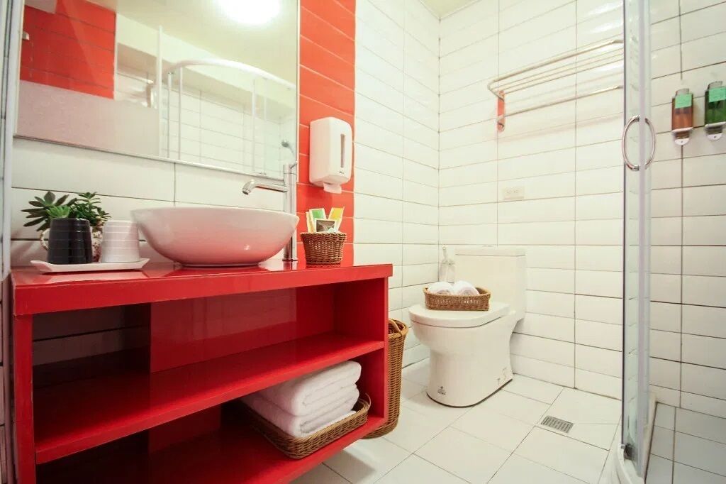 A modern bathroom with a red vanity, white tiles, a large mirror, a white toilet, and a glass shower enclosure.