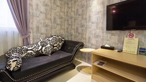 Una habitación con un sofá oscuro, un televisor de pantalla plana montado en la pared, una mesita redonda pequeña y un estante de madera con un letrero.