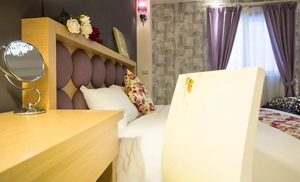A hotel room with a wooden desk, a round mirror, a bed with white bedding, a decorative headboard, and a window with curtains.