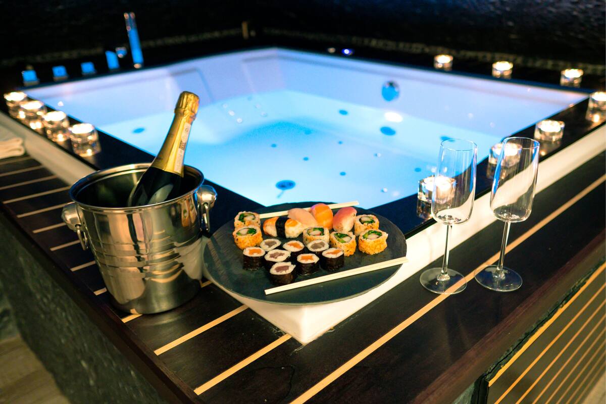 A hot tub with a champagne bottle, sushi, and wine glasses on a wooden deck.