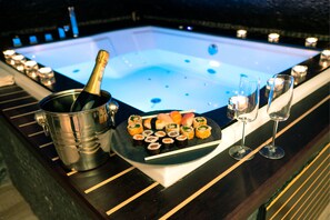 Un jacuzzi con una botella de champán, sushi y copas de vino sobre una terraza de madera.