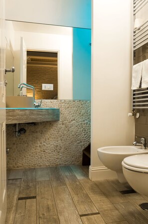 Un baño con piso de madera, lavamanos de vidrio, inodoro blanco y pared azul.