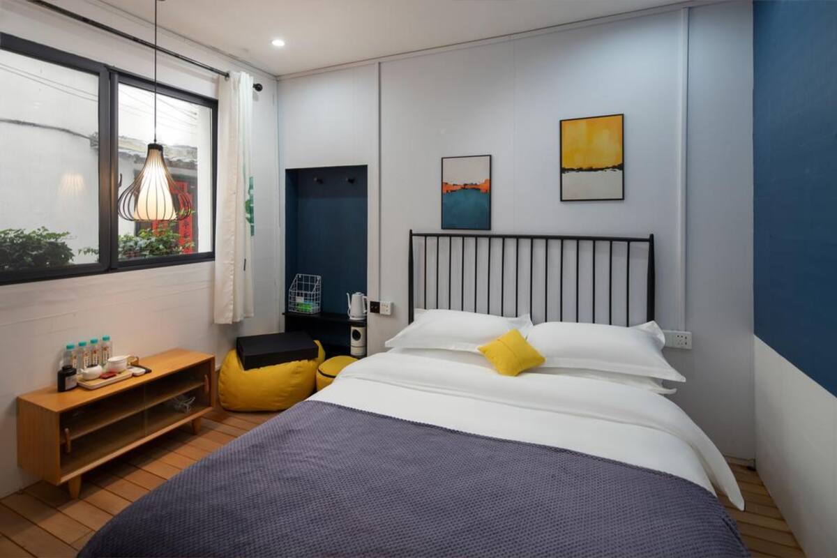Ein modernes Schlafzimmer mit einem Bett, einem Nachttisch mit Pflegeprodukten, einem Fenster und einem Kunstwerk an der Wand.