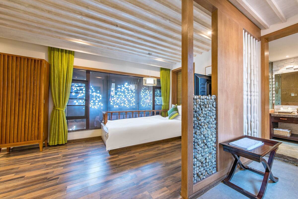 A hotel room with a large bed, wooden flooring, a bathroom with a sink and mirror, and a view through a window with curtains.