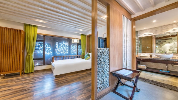 Habitación de hotel con una cama grande, suelo de madera, baño con lavamanos y espejo, y vistas a través de una ventana con cortinas.