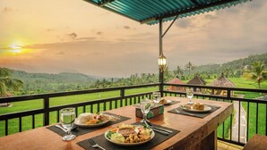 Tavola imbandita con piatti di cibo, bicchieri e una lampada, con vista su un paesaggio rigoglioso e un tramonto.