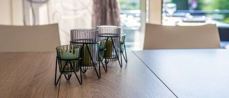 Een eettafel met vier kandelaars van draad en uitzicht op een balkon met een hekwerk en groen.