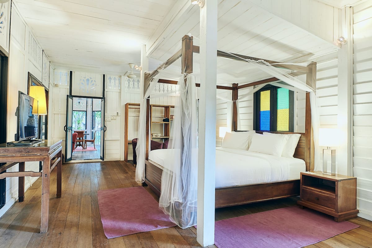 Un dormitorio con cama con dosel, suelo de madera y vistas a una habitación colorida a través de una puerta.
