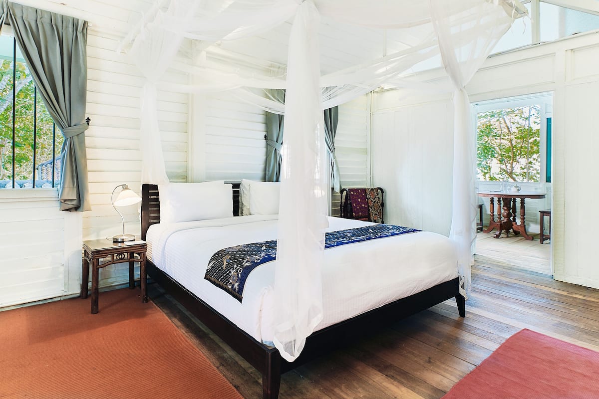 Una cama con dosel y cuatro columnas, ropa de cama blanca, una mesita de noche de madera con lámpara y vistas al exterior a través de ventanas amplias.