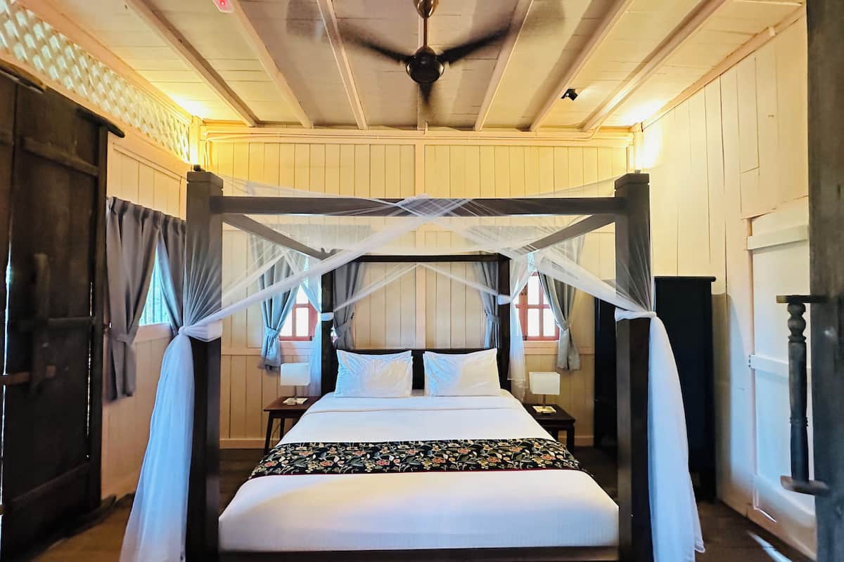 Una cama con dosel y cuatro columnas, con un edredón estampado con motivos florales, un ventilador de techo y una mesita de noche con lámpara.