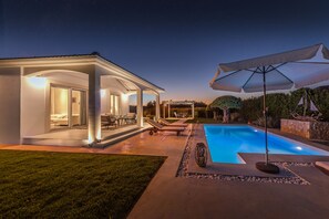 A modern outdoor pool area with a covered seating area, lounge chairs, and a well-lit patio. - Villa Evanna (Corfu)