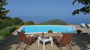 Un espace aménagé au bord de la piscine, avec deux chaises en bois et une table, d’où l’on peut admirer une piscine aux eaux d’un bleu limpide et un paysage montagneux.