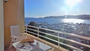 A balcony with a table, a chair, and a view of a coastal town and sea.