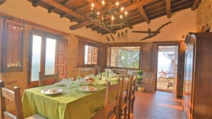 Un espace repas avec une longue table dressée pour le repas, des poutres en bois au plafond et une vue sur l’extérieur par la porte ouverte.
