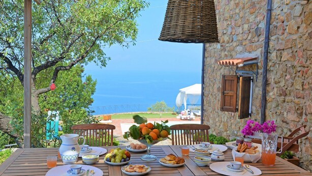 Une table dressée pour le petit-déjeuner, avec vue sur la mer, un bâtiment en pierre et une pergola.