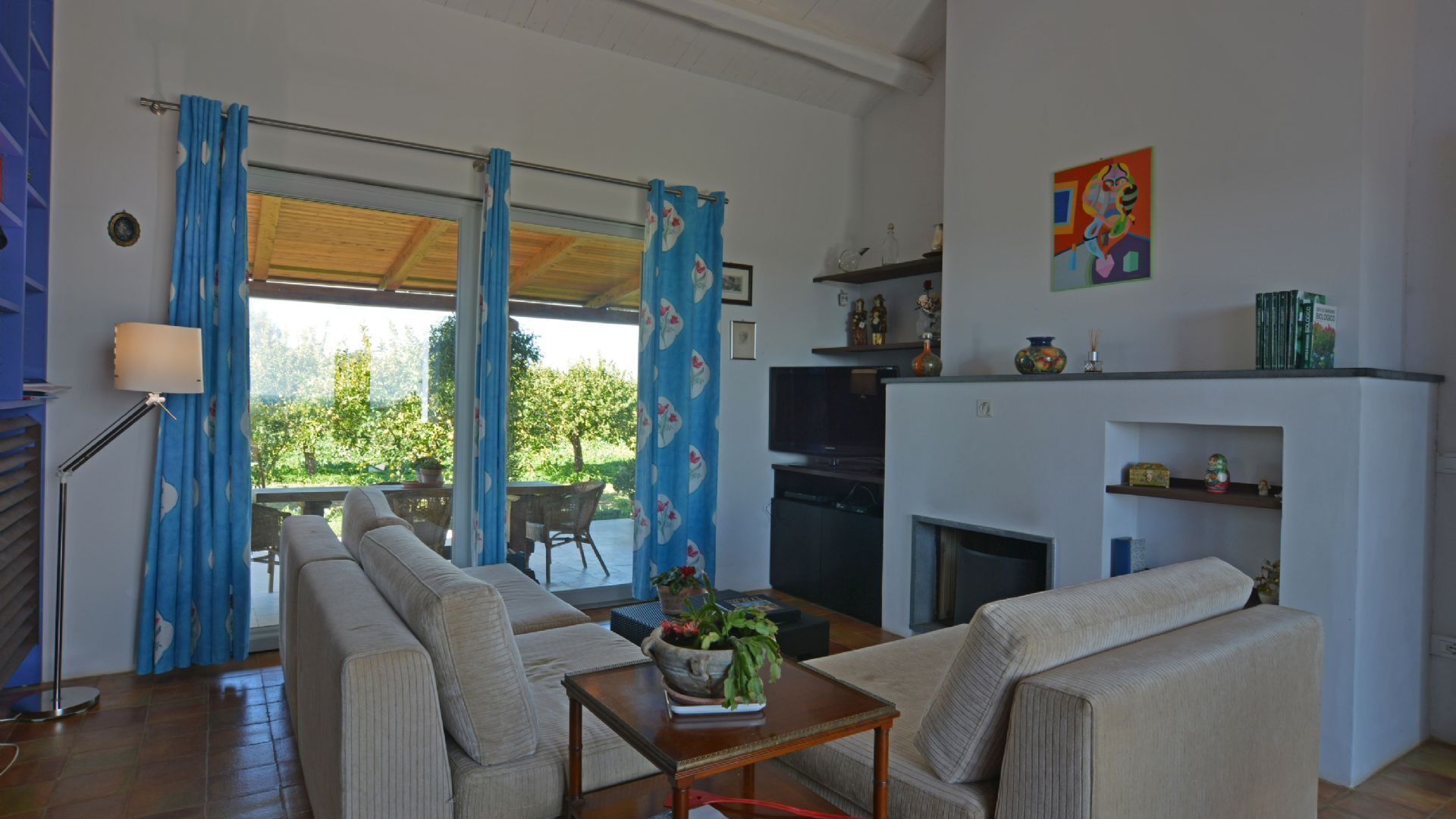 Een woonkamer met een bank, een salontafel met een plant, een televisie en een open haard. Er is een balkon met blauwe gordijnen en uitzicht op groen.