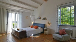 Une chambre à coucher avec un lit, une table de chevet, une chaise, une fenêtre et une vue sur la verdure à l’extérieur.