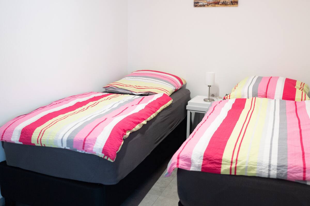 Une petite chambre propre, avec deux lits simples, une table de chevet et une lampe fixée au mur.