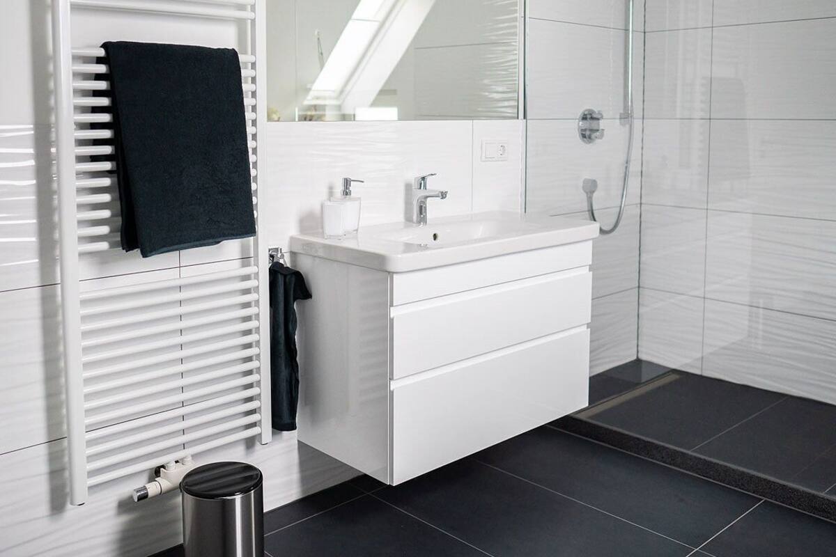 Une salle de bain moderne avec un meuble-lavabo blanc, un grand miroir, une douche équipée d’un pommeau de douche à main et un radiateur.
