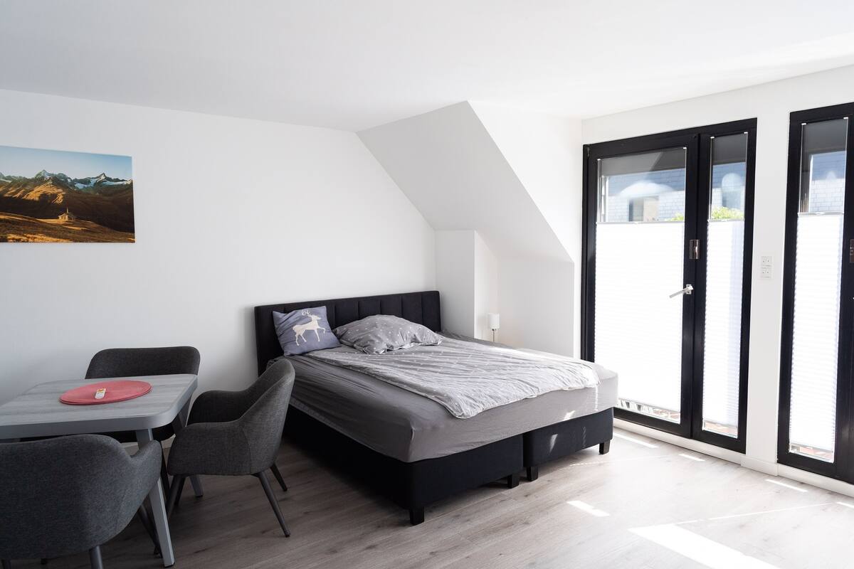 Une chambre moderne avec un lit, un bureau et des chaises. Une grande fenêtre offre une vue sur les bâtiments.