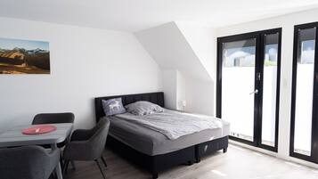 Ein modernes Schlafzimmer mit einem Bett, einem Schreibtisch und Stühlen. Ein großes Fenster bietet Blick auf Gebäude.