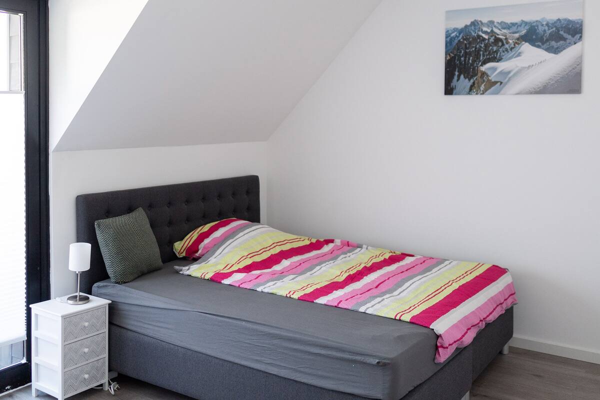 Une chambre moderne avec un lit gris, une couverture à rayures, une tête de lit grise, une table de chevet avec une lampe et un tableau représentant un paysage de montagne accroché au mur.