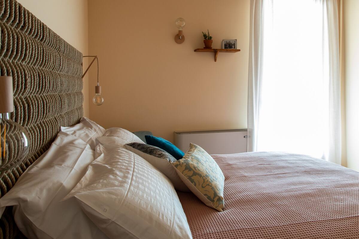 A neatly made bed with white and patterned pillows, a textured headboard, and a small shelf with a plant and picture frame.