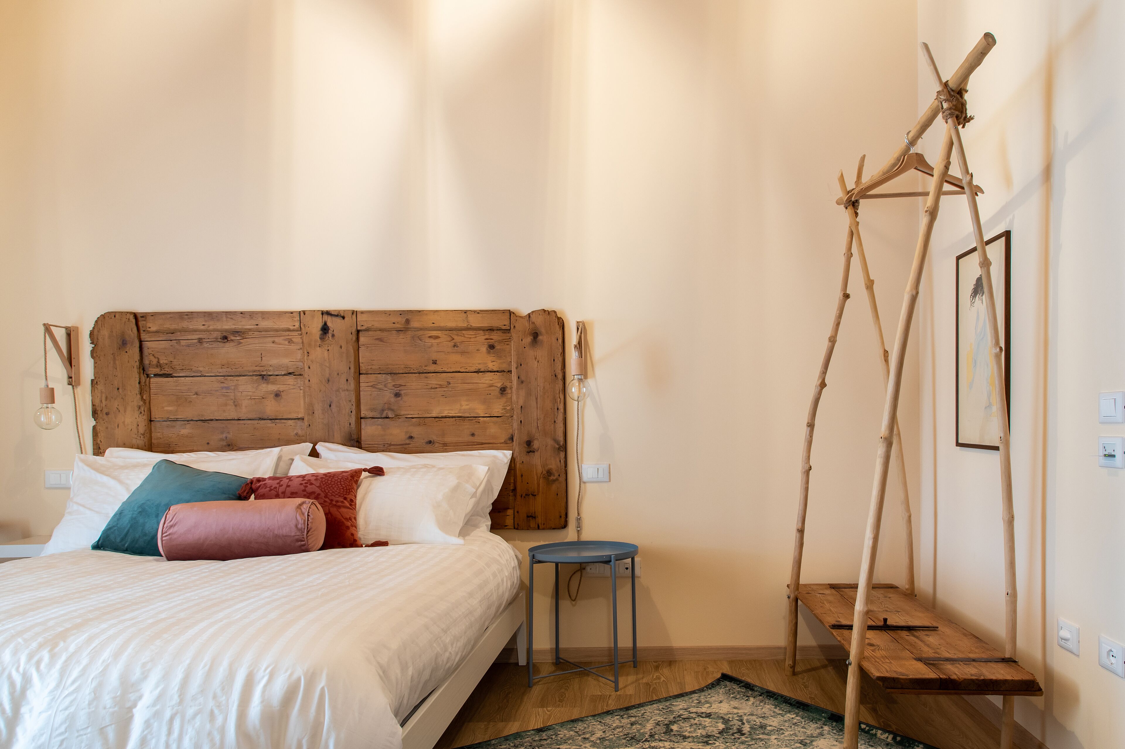 Quarto com cabeceira de madeira, cama com almofadas brancas e coloridas, um banquinho azul pequeno e uma cadeira de balanço de madeira.