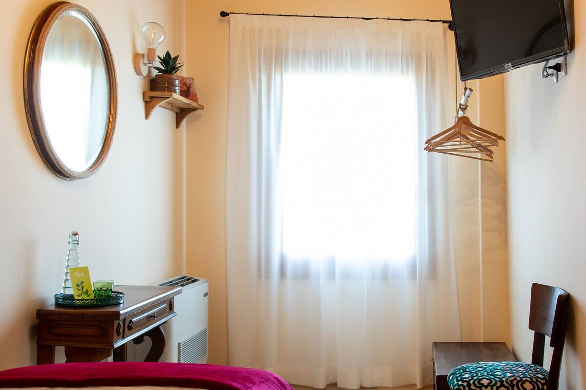 A bedroom with a bed, a desk, a chair, a TV, and a window with curtains.