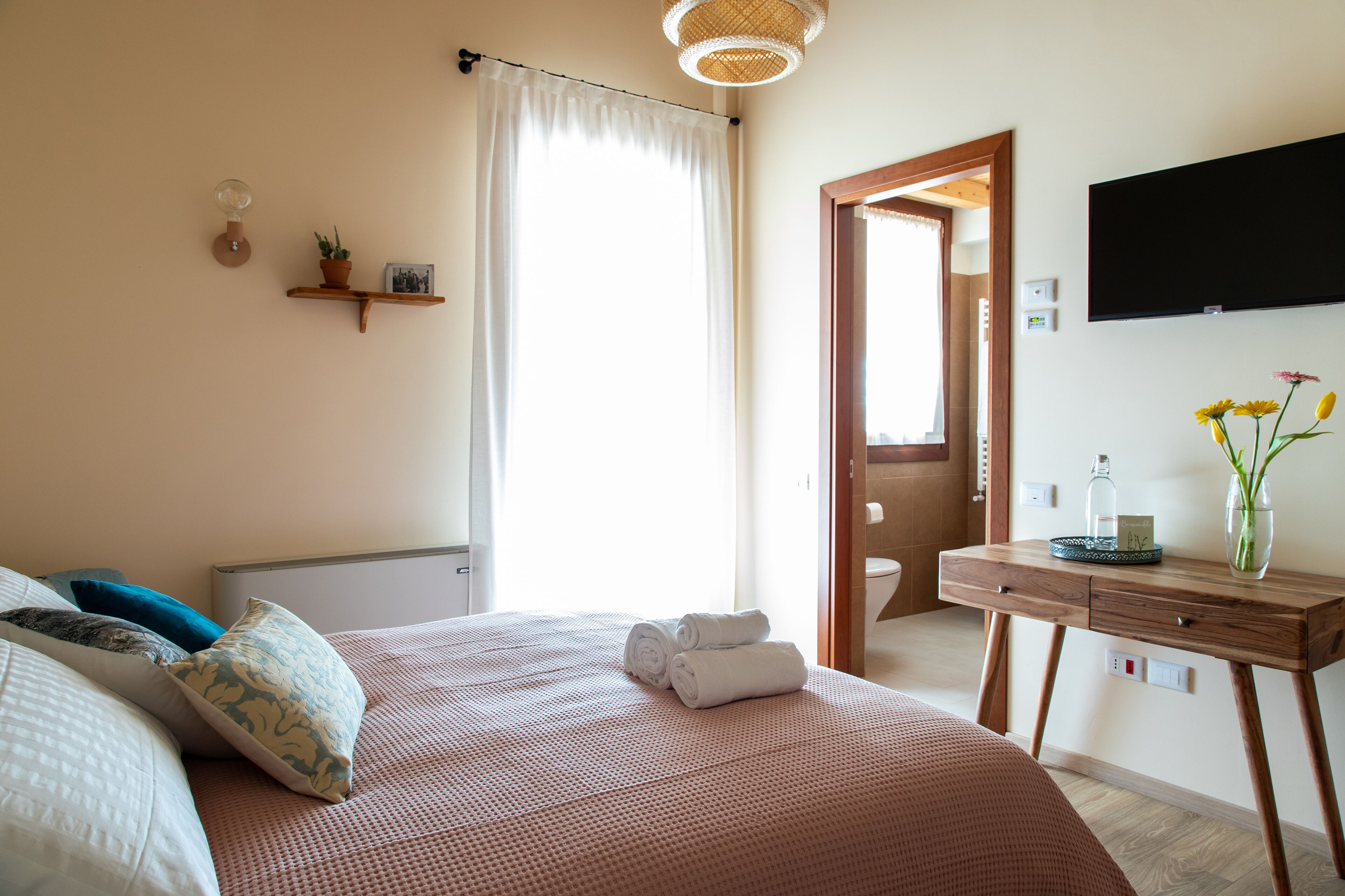 Une chambre à coucher comprenant un lit, des oreillers, une table de chevet avec un vase, un téléviseur à écran plat et une porte donnant accès à une salle de bain.