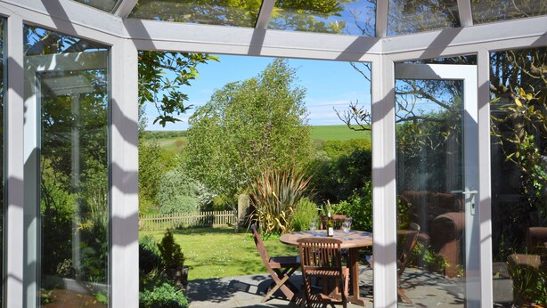 Une véranda dotée d’une porte vitrée, une table ronde avec des chaises, et une vue sur un jardin avec une clôture en bois et des arbres.
