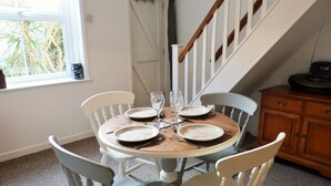 Un coin repas avec une table ronde dressée pour quatre personnes, des chaises blanches, un buffet en bois et un escalier en arrière-plan.