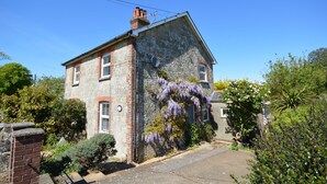 Une maison en pierre à deux étages, dotée d’une cheminée en briques, d’une petite fenêtre et d’un jardin où poussent des plantes à fleurs violettes.