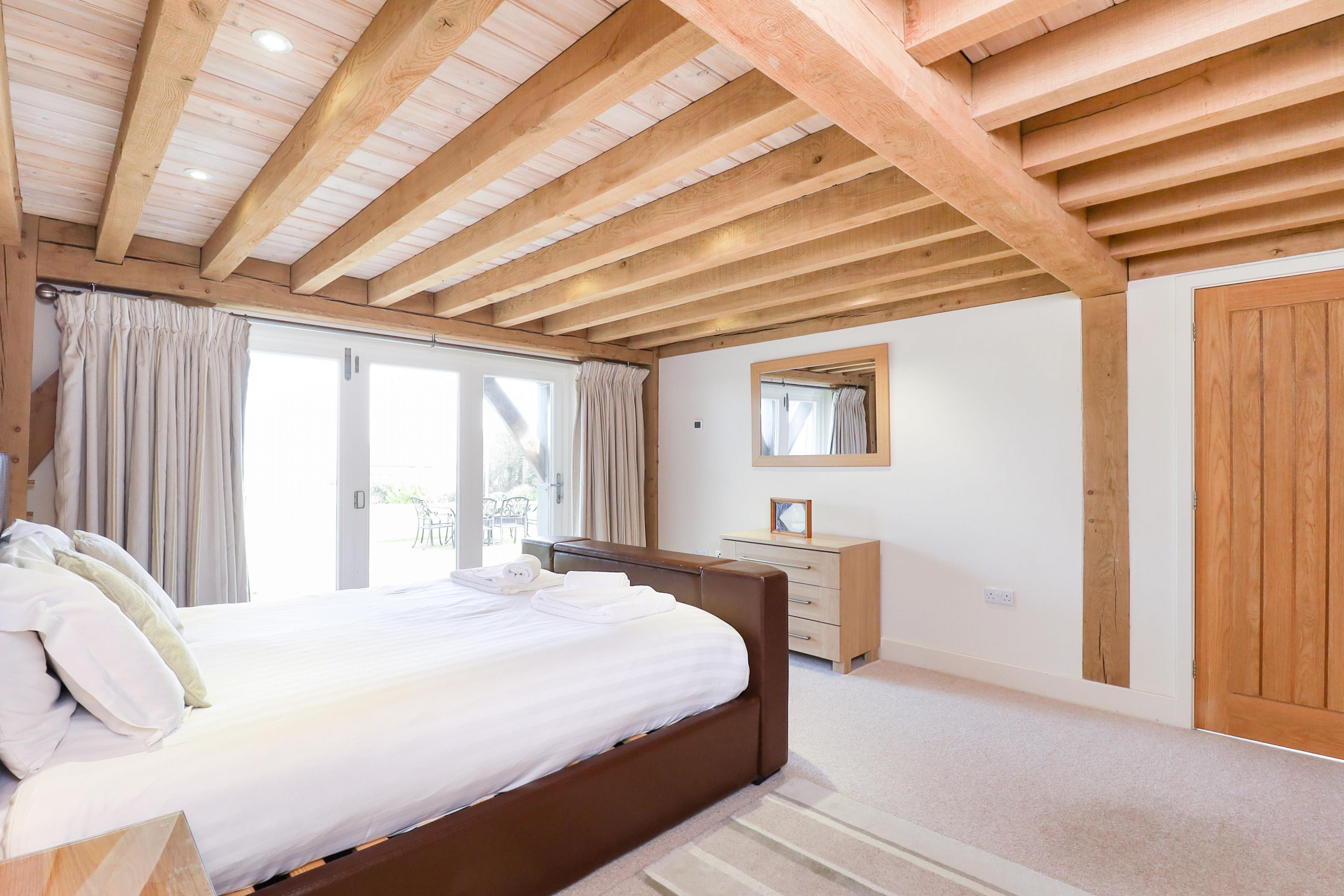 Ein Schlafzimmer mit Holzbalkendecke, einem Bett mit weißer Bettwäsche, einer hölzernen Kommode und einem großen Spiegel an der Wand.