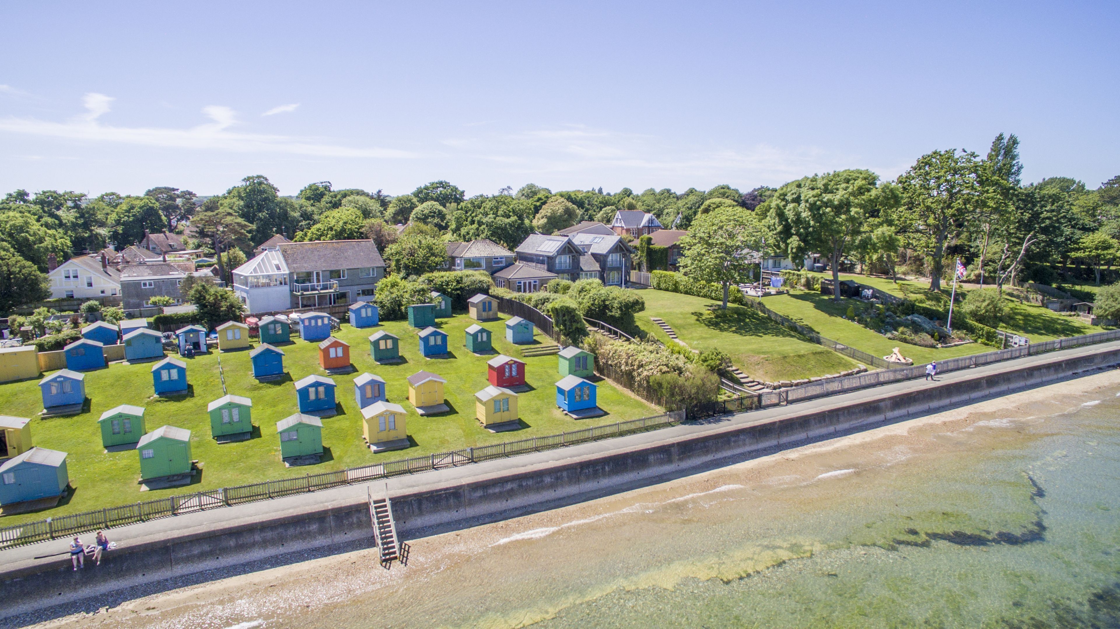 A coastal area with colorful beach huts arranged in rows on a grassy field, a concrete walkway beside the water, and residential buildings in the background.