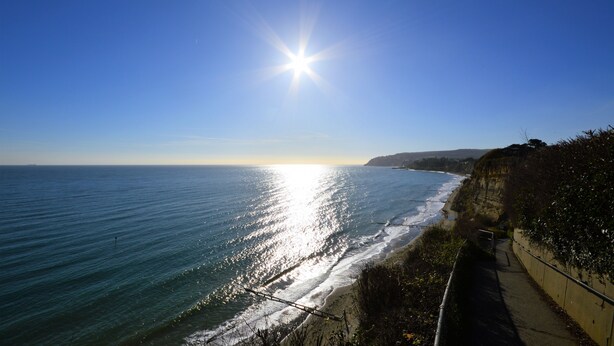 Vista costera con cielo despejado, el sol reflejándose en el agua y un paseo por el borde del acantilado.