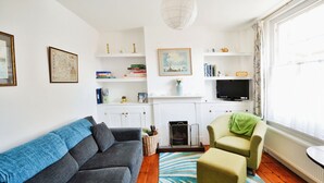 A living room with a grey sofa, a green armchair, a blue and white striped rug, a fireplace, and a television. - Admirals Cottage (Yarmouth)