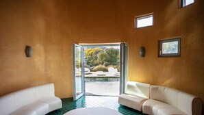Una spa con un divano bianco curvo, un tavolino con un libro e una porta vetrata che dà su una zona esterna con piscina e vegetazione.