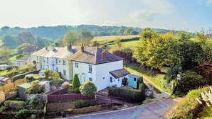 A two-story house with a white facade and multiple chimneys, surrounded by lush greenery and a hilly landscape. - Charnwood Cottage (Sandown)