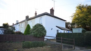 A two-story residential house with a white exterior, a brick chimney, and a small garden area with a hedge and a tall, narrow bush. - Charnwood Cottage (Sandown)