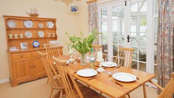 A dining room with a wooden table set for four, surrounded by wooden chairs. A wooden cabinet with various dishes and a ship decoration is visible in the background.