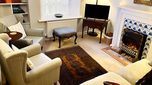 A living room with a sofa, armchair, and ottoman, a fireplace, a television, and a bookshelf with books and decorative items. - Grove Cottage (Yarmouth)