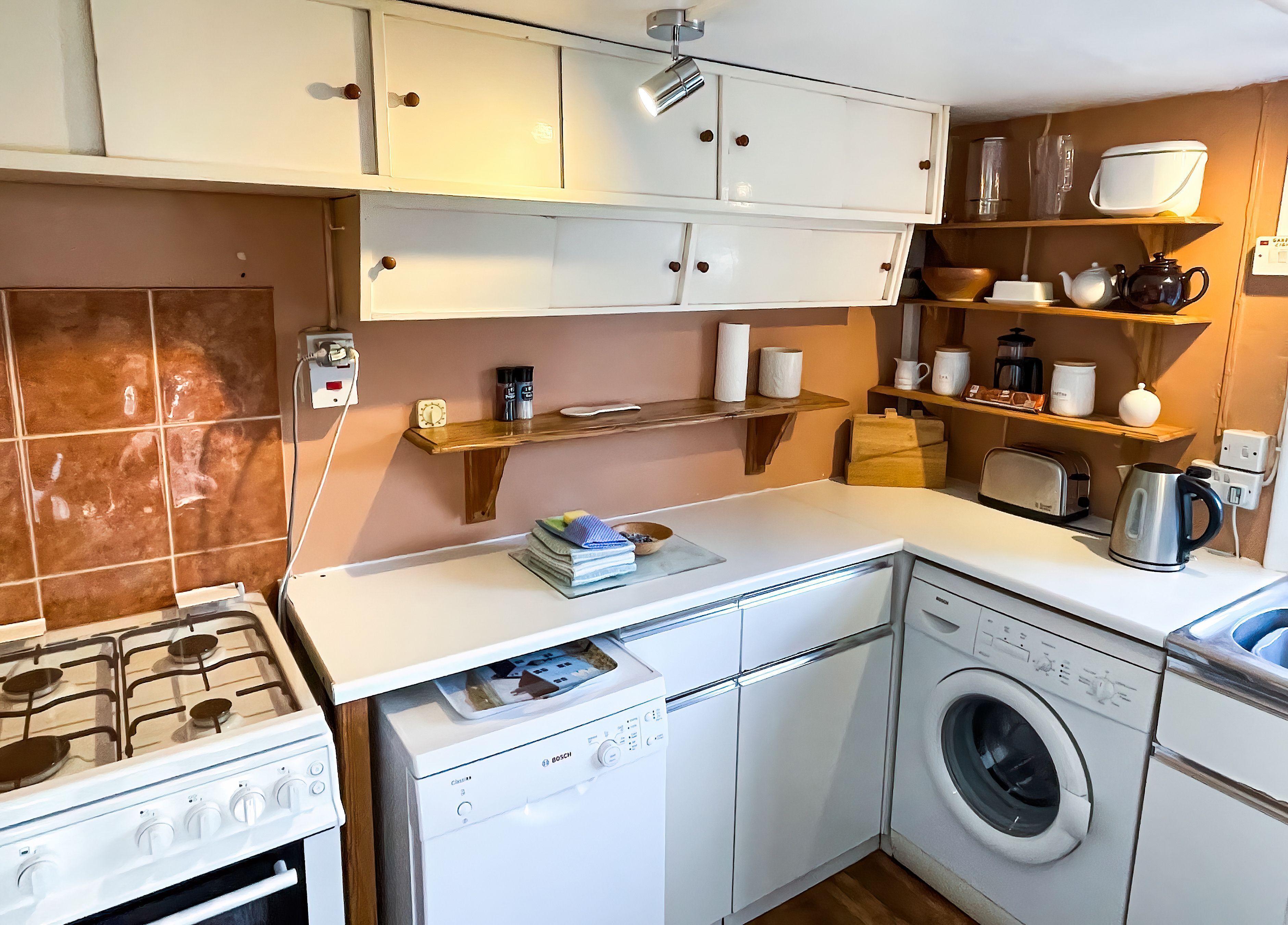 A compact kitchen with a Bosch washing machine, a gas stove, and wooden shelves above the countertop.